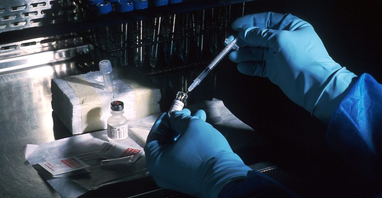 Gloved hands working with vaccine vials and a syringe in a fume hood
