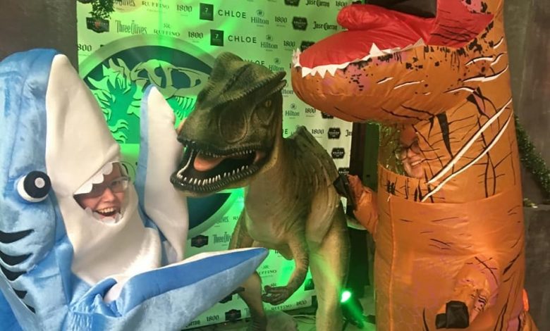 a person in a left shark costume, a person in an inflatable trex and a model dinosaur