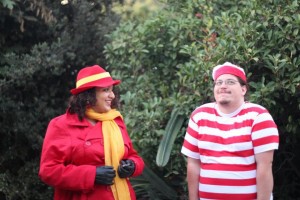 on left, a woman is smiling and dressed in a Carmen Sandiego costume. on right, a man dressed up as Where's Waldo? is shrugging with a smile.