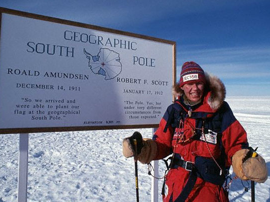 Women in cold weather gear beside sign for South Pole